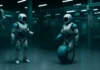 Två futuristiska robotar arbetar i fabrik – en med hjul i stället för ben – i en högteknologisk AI-miljö med turkosa ljuseffekter